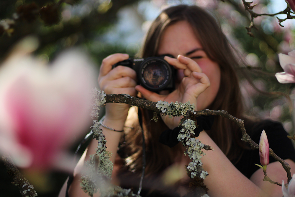 Portrait d'une jeune femme avec un appareil photo devant le visage, entourée par des fleurs de cerisier