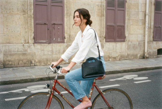 Une femme en jean et chemise blanche sur un vélo de ville, elle porte le sac LUA N°1.